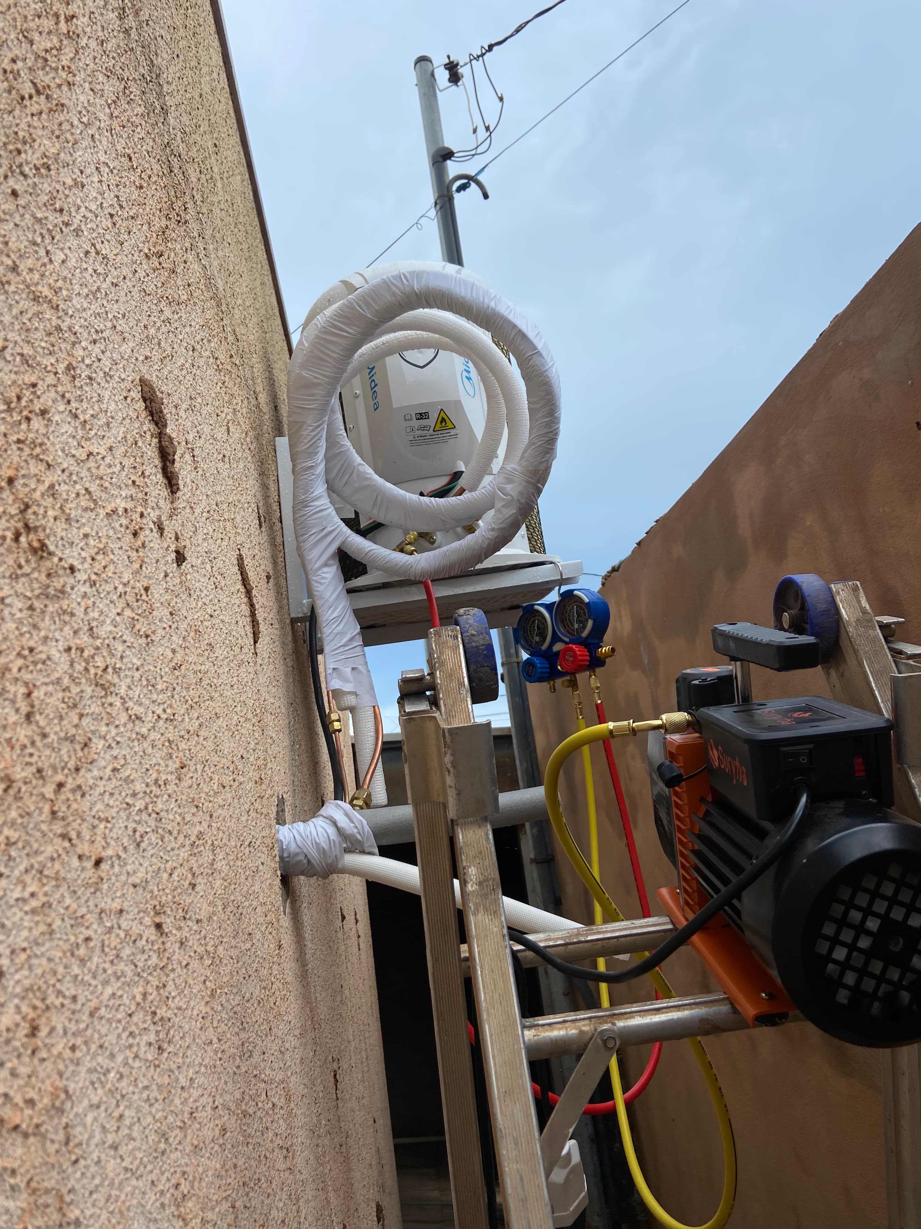 Instalação de ar-condicionado com tubos brancos isolados e manômetro em uma parede externa.