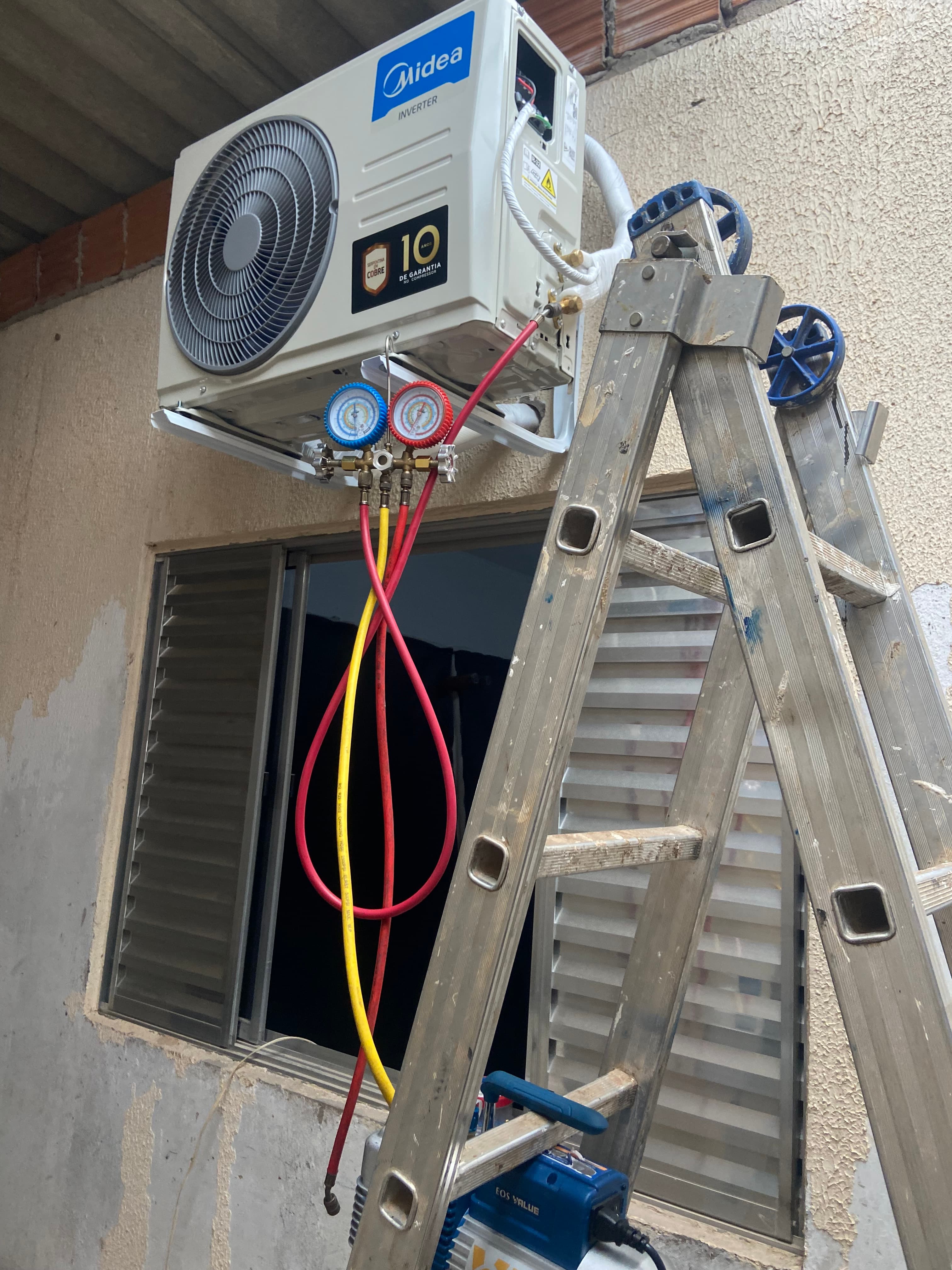 Ar-condicionado Midea em manutenção com manômetro e mangueiras coloridas sob uma escada de alumínio.