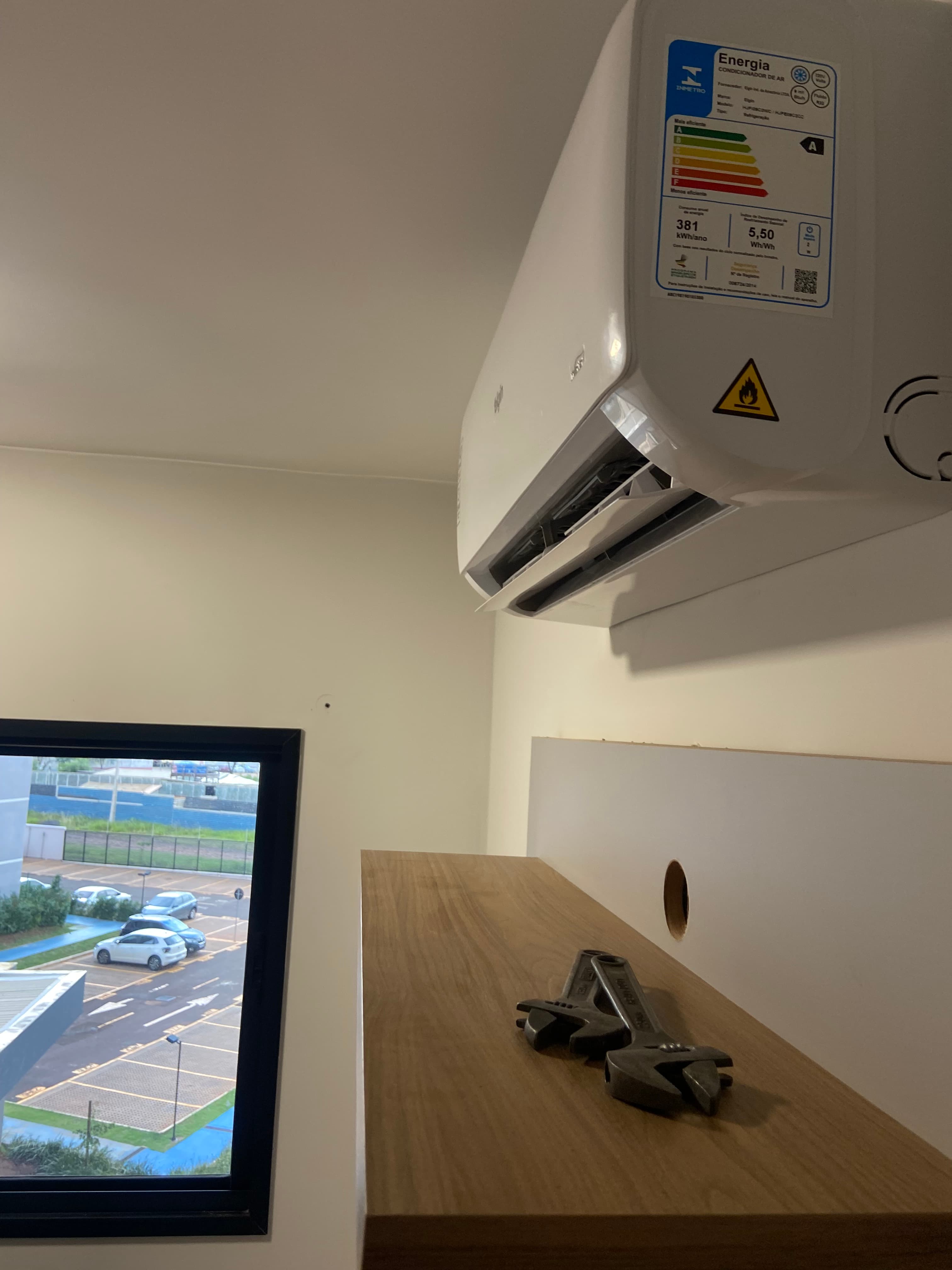 Ar condicionado branco na parede, prateleira de madeira com ferramentas e vista para estacionamento.