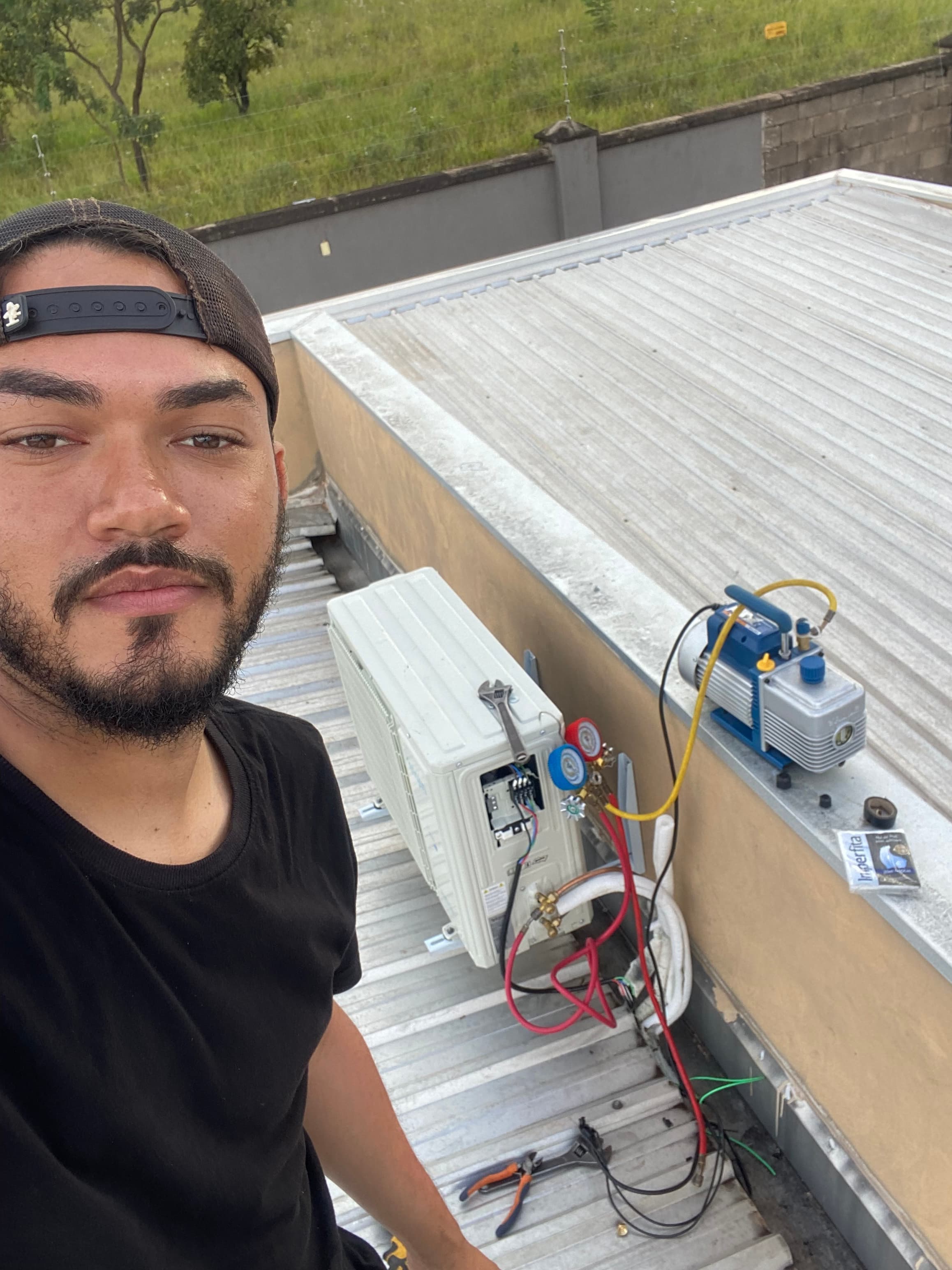Técnico tira selfie durante manutenção de ar-condicionado em telhado metálico com bomba de vácuo.