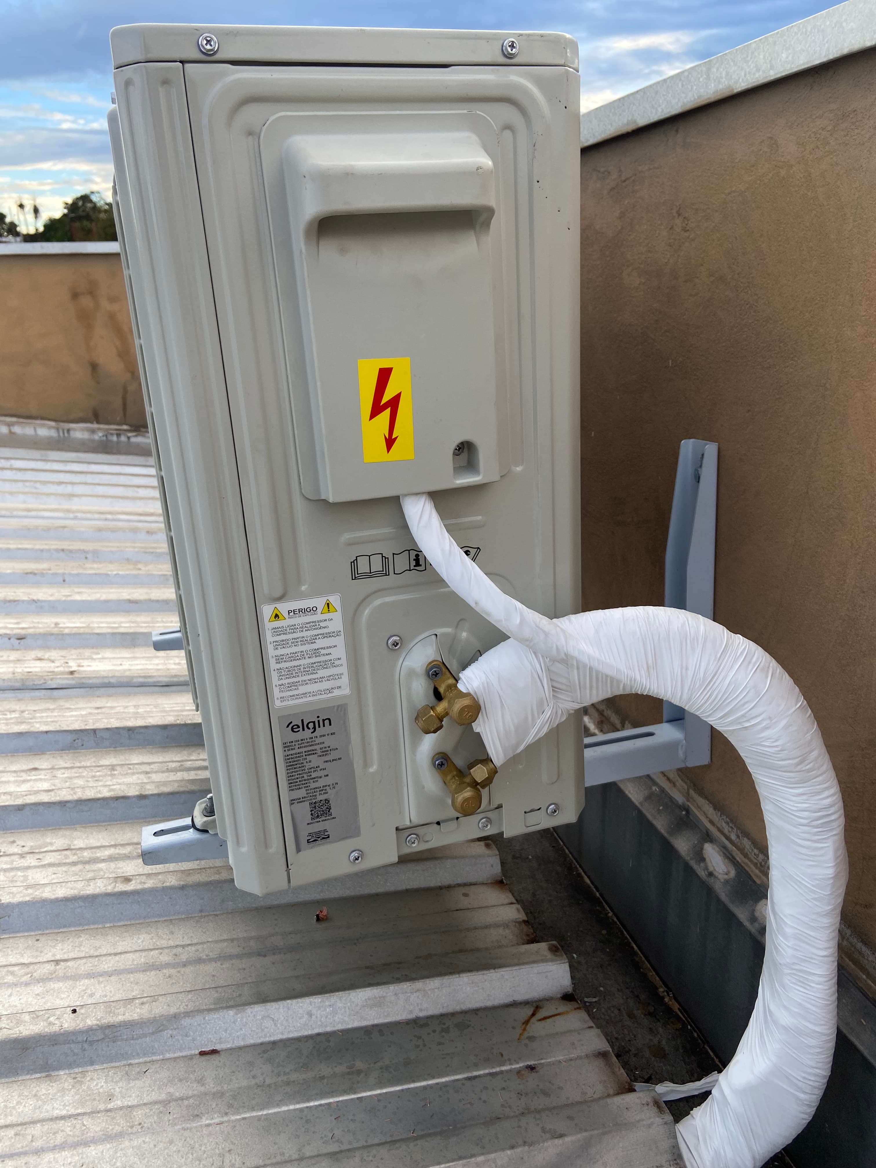 Unidade externa de ar-condicionado Elgin com tubulação isolada branca e etiqueta de aviso elétrico.