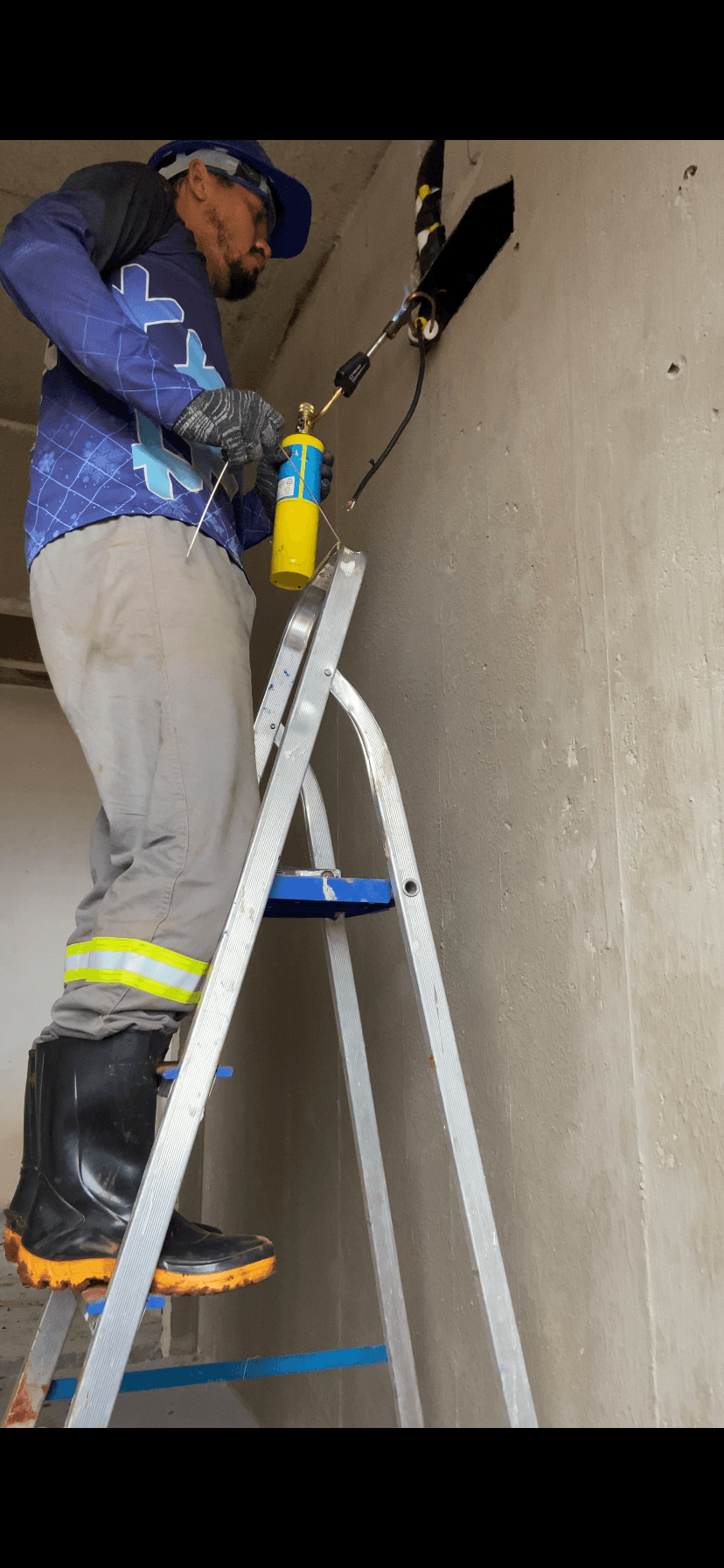 Trabalhador em escada usa maçarico para soldar tubos em uma parede de concreto inacabada.