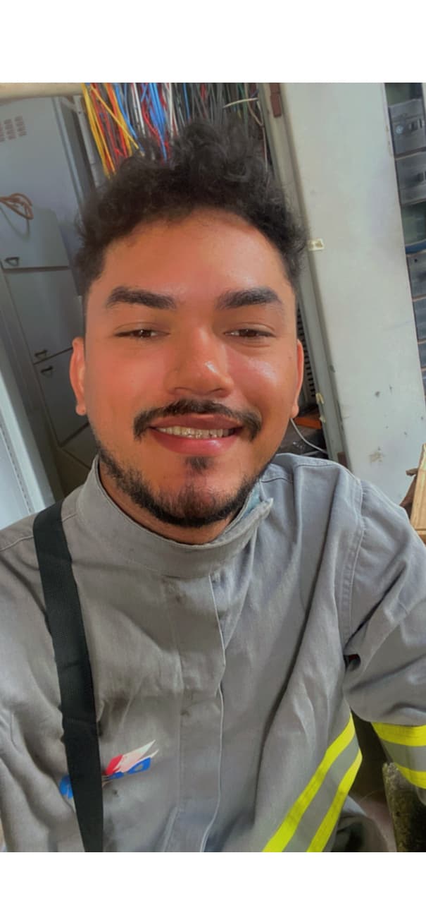 Homem jovem sorridente com barba, vestindo uniforme cinza com faixas refletivas em ambiente técnico.